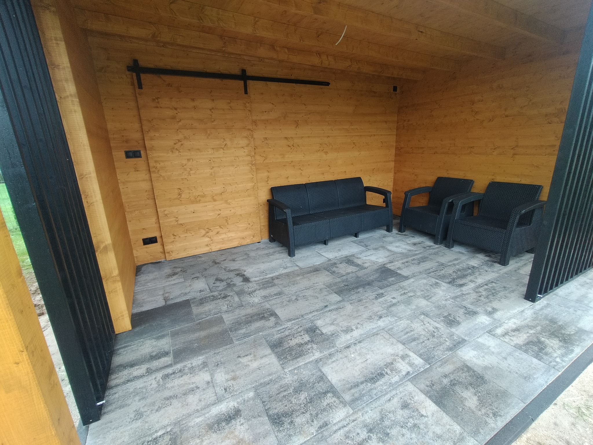 Zadaszony taras z drewnianą ścianą, przesuwanymi drzwiami, szarą kostką brukową i czarnymi meblami ogrodowymi z technorattanu: sofa i dwa fotele.