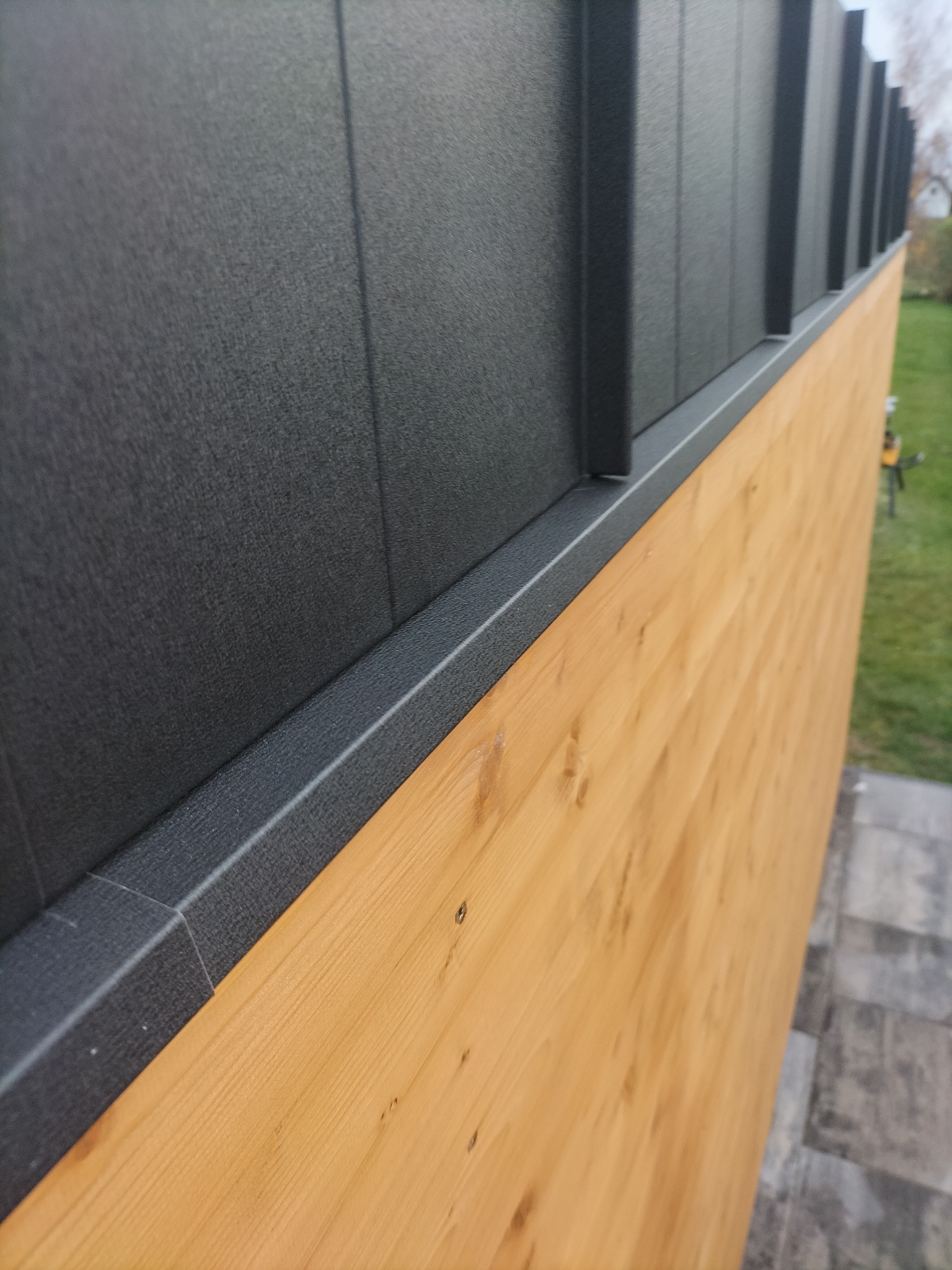 Szczegółowe ujęcie elewacji budynku z drewnianą boazerią i czarnymi panelami elewacyjnymi, widoczne śruby mocujące, zieleń w tle.