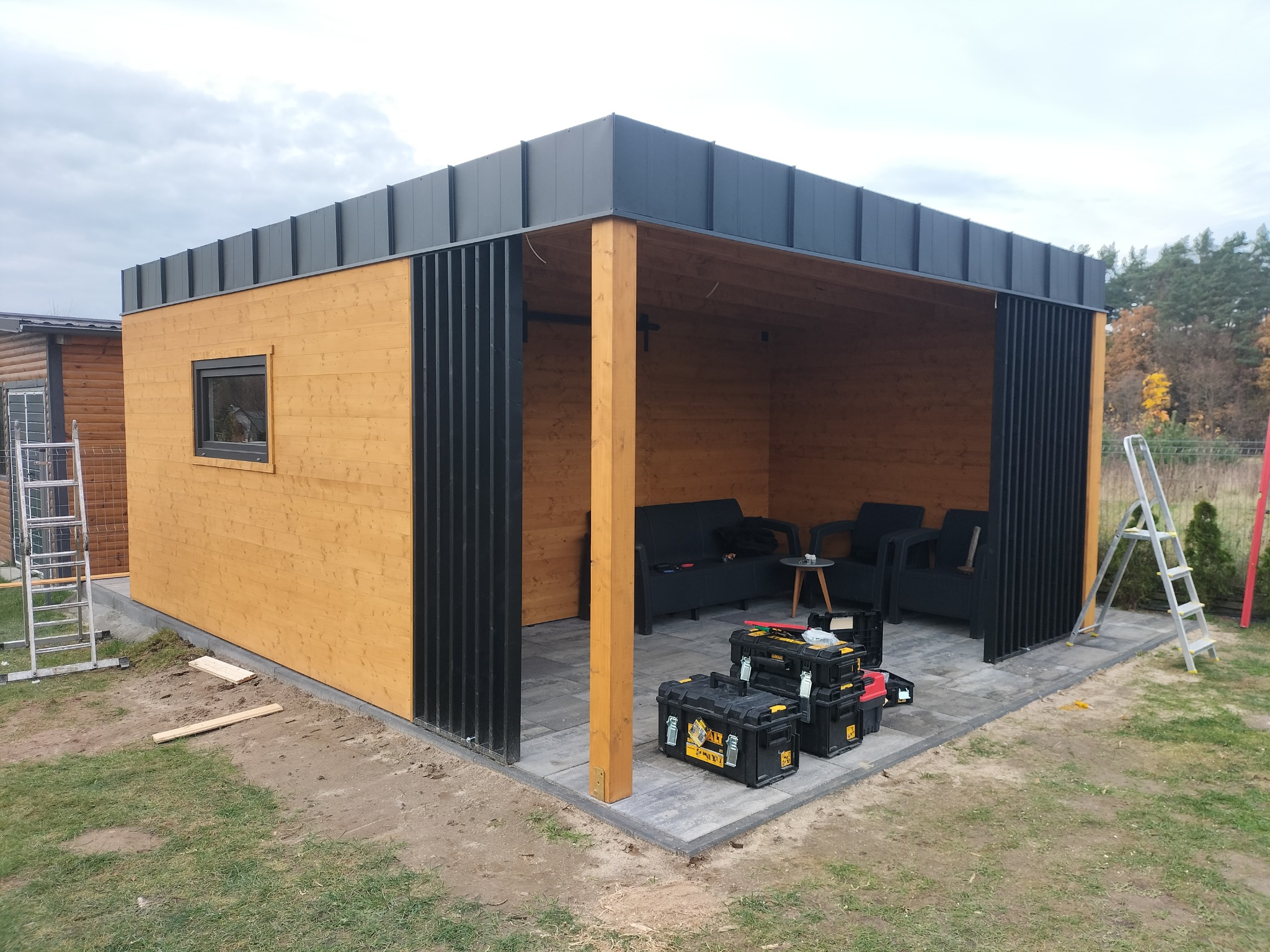 Drewniana altana z czarnymi elementami wykończeniowymi, częściowo zamknięta, z meblami ogrodowymi wewnątrz, skrzynki z narzędziami na zewnątrz, widoczne drabiny i przygotowany teren.