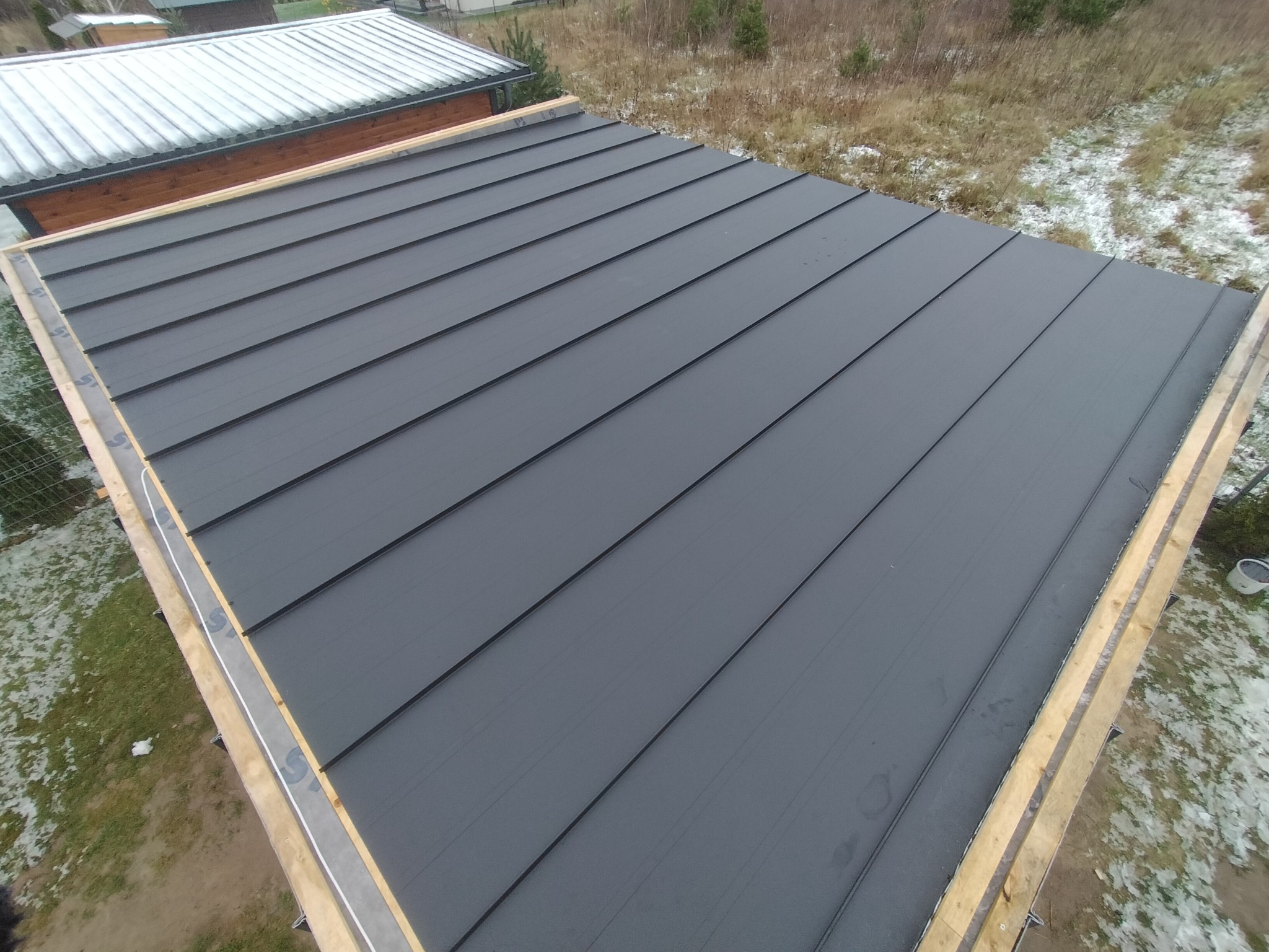 Widok z góry na nowy, grafitowy dach z blachy na drewnianej konstrukcji, częściowo pokryty śniegiem, w tle trawa i krzewy.