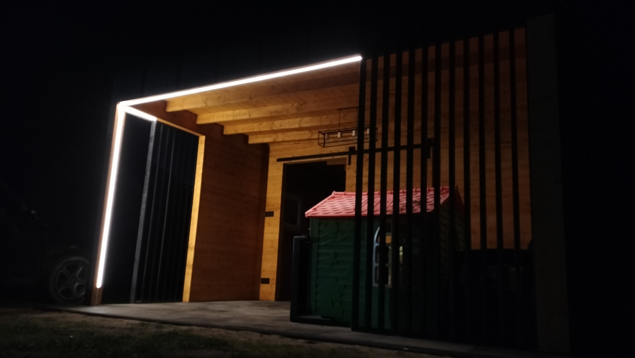 Nowoczesna wiata garażowa z drewnianą konstrukcją, oświetleniem LED i elementami z pionowych, czarnych listew, widziana nocą zza ogrodzenia.