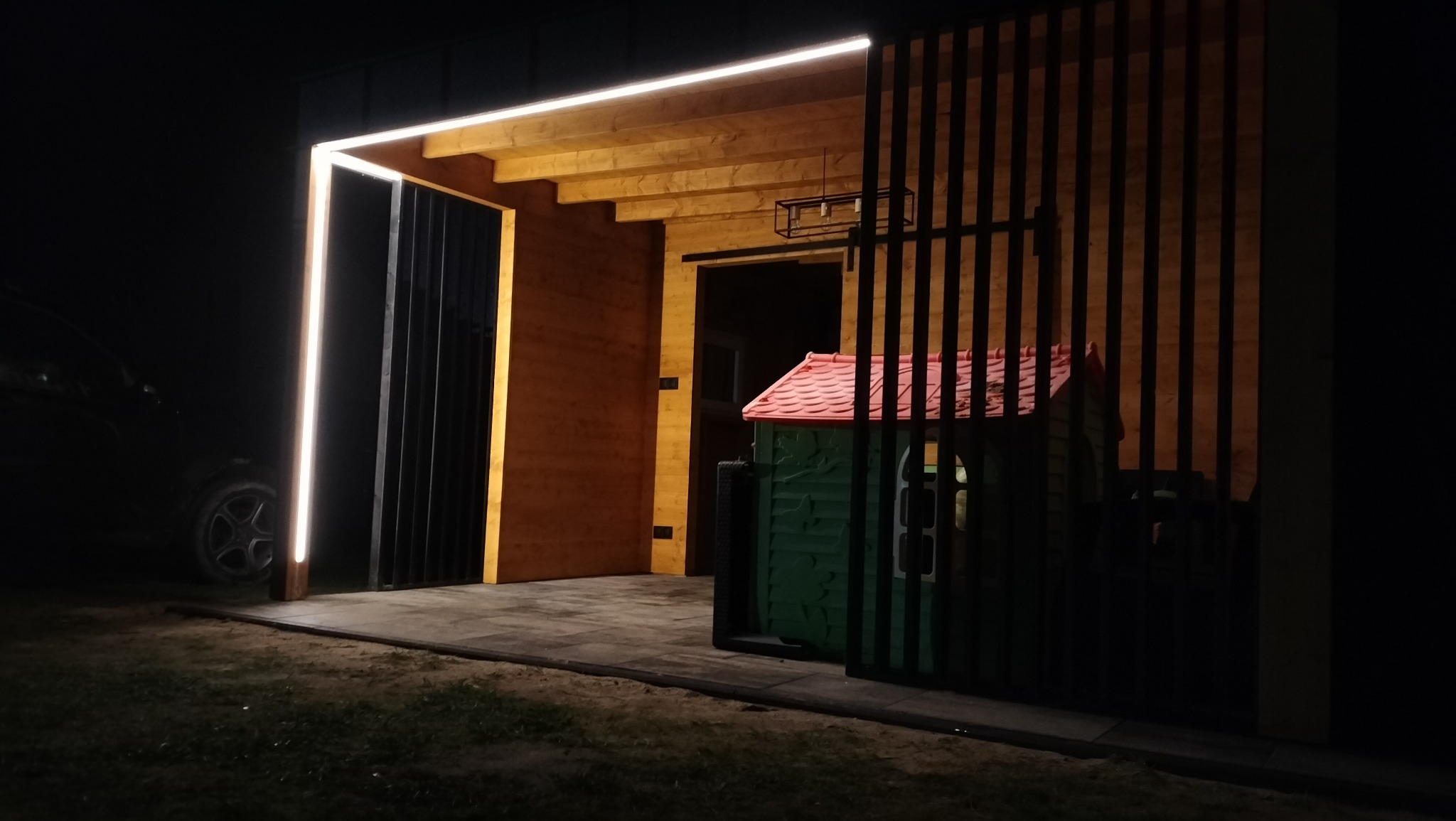 Nowoczesna altana z drewnianym wykończeniem i oświetleniem LED w Pucku, widoczna przez pionowe elementy ogrodzenia, z małym domkiem dla dzieci w tle.