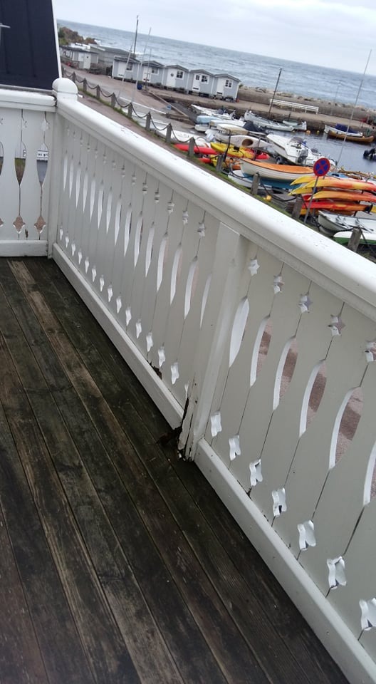 Drewniany balkon z widokiem na przystań z łodziami, uszkodzona biała balustrada z dekoracyjnymi wycięciami, ciemne deski podłogowe.