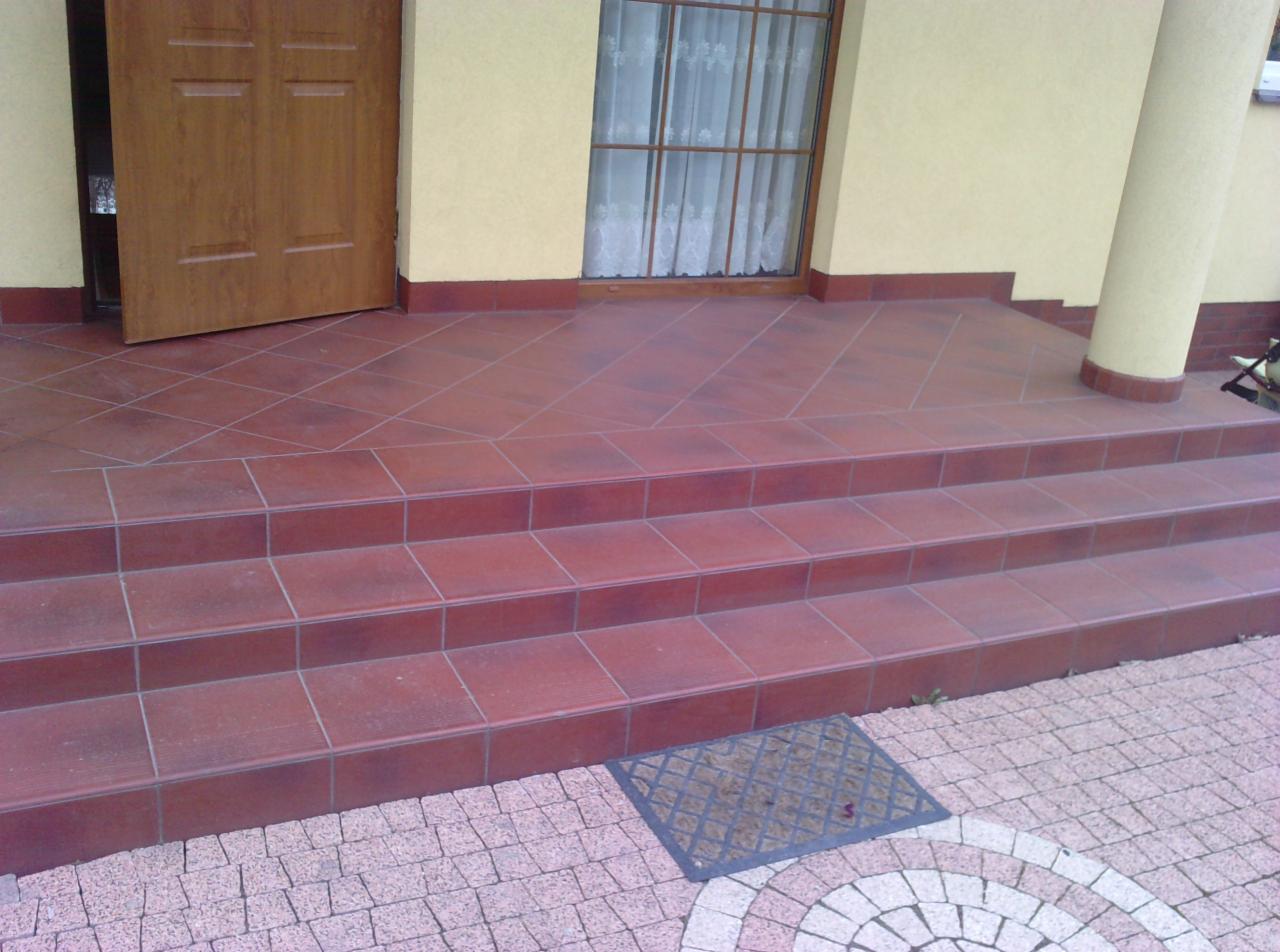 Kafelkowane schody wejściowe z otwartymi drzwiami i widokiem na kostkę brukową ułożoną w dekoracyjny wzór koła. Kolumna przy wejściu.