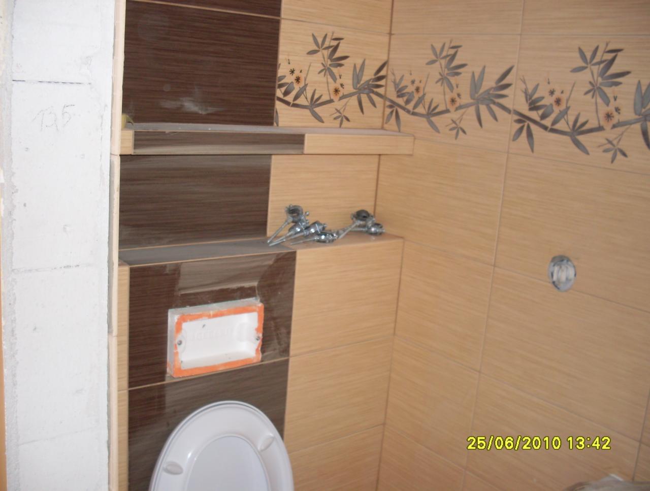 Wykończona łazienka z beżowo-brązowymi płytkami, dekoracyjnym motywem roślinnym i półkami. Widoczne elementy instalacji wodnej i fragment toalety.