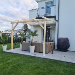 Piotr Wicherek - Surowa konstrukcja z jasnego drewna, prawdopodobnie w trakcie montażu, tworząca zadaszenie nad tarasem z meblami ogrodowymi i palmami w donicach, na tle białego domu z balkonem.