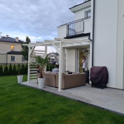 Piotr Wicherek - Biała pergola z drewnianą konstrukcją osłaniająca rattanowy narożnik na szarym tarasie, z palmami w donicach i grillem pod pokrowcem, na tle zadbanego trawnika i nowoczesnego domu.