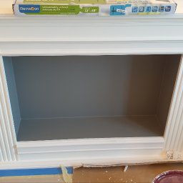 Piotr Wicherek - Biały, klasyczny kominek w trakcie malowania na szaro wewnątrz, widoczne puszki z farbą, taśma malarska i karton zabezpieczający podłogę, na górze opakowanie uchwytu ściennego do TV.