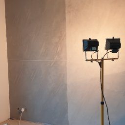 Piotr Wicherek - Ściana po dekoracyjnym malowaniu z efektem betonu, oświetlona dwoma halogenami na statywie, widoczne narzędzia malarskie i zabezpieczenia podłogi.