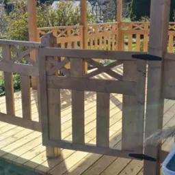 Drewniana altana z ażurową balustradą i furtką, z widokiem na ogród i dom w tle.