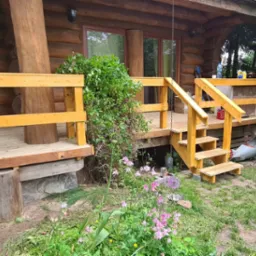 Drewniany taras przy domu z bali, z żółtymi barierkami i schodami. Elementy konstrukcyjne oparte na kamieniach. Bujna roślinność w tle i na pierwszym planie.
