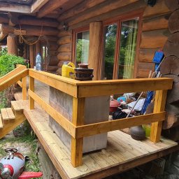 Piotr Wicherek - Drewniany taras przy domu z bali, widoczne schody i balustrada. Na tarasie ustawione donice i inne przedmioty. W tle drzewa i krzewy.