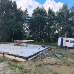 Betonowa płyta fundamentowa z izolacją, przygotowana pod budowę. W tle zieleń drzew, przyczepa kempingowa i biały samochód dostawczy.
