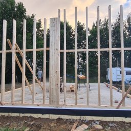 Piotr Wicherek - Surowa konstrukcja drewniana budynku w trakcie budowy, widoczne słupy i belki, na tle zalesionego terenu. W tle przyczepa kempingowa i samochód dostawczy.