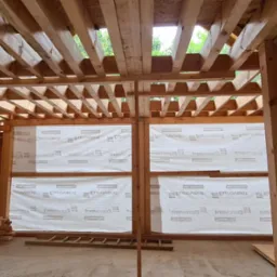 Konstrukcja drewniana w trakcie budowy: szkielet ścian z membraną, widoczna więźba dachowa, częściowo wykonane schody. Wnętrze w stanie surowym, przygotowane do dalszych prac.