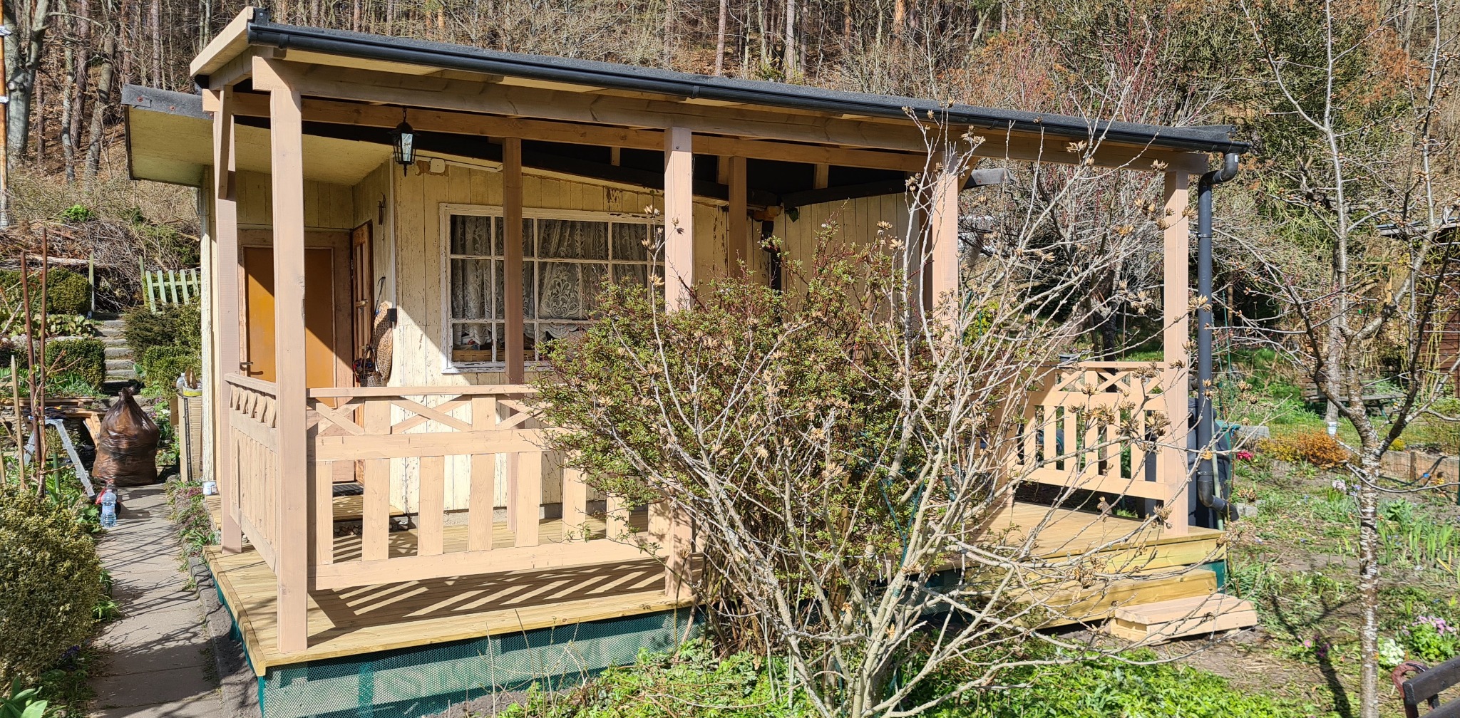 Drewniana konstrukcja z zadaszeniem i balustradą, prawdopodobnie weranda lub mała altana, z widocznymi śladami użytkowania i otoczona zielenią w ogrodzie.