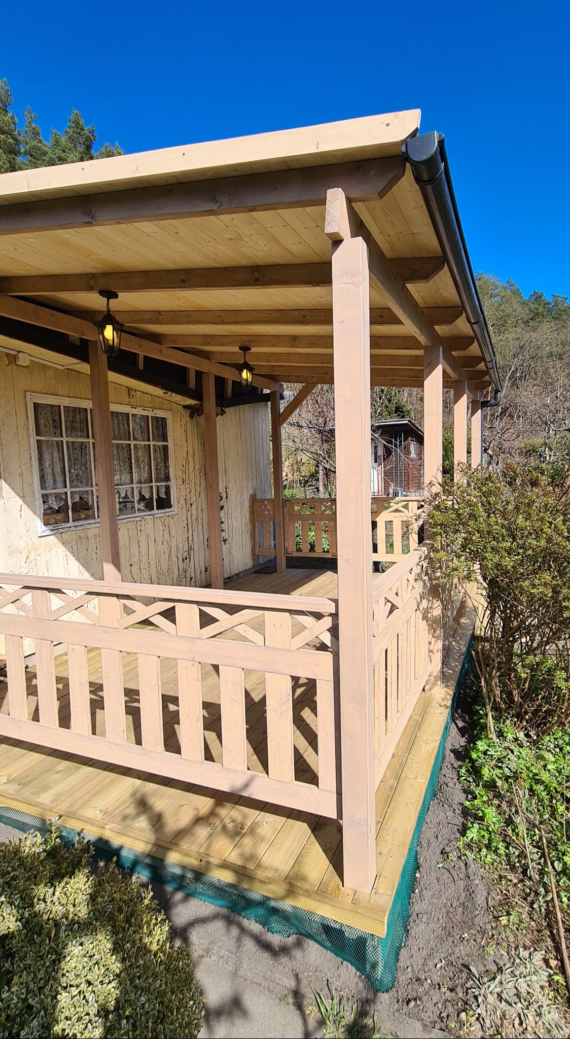 Zadaszony taras z drewnianą podłogą i balustradą w kolorze beżowym, oświetlony dwoma wiszącymi lampami w stylu retro, przylegający do budynku z oknami, z widocznymi elementami ogrodu i siatką...