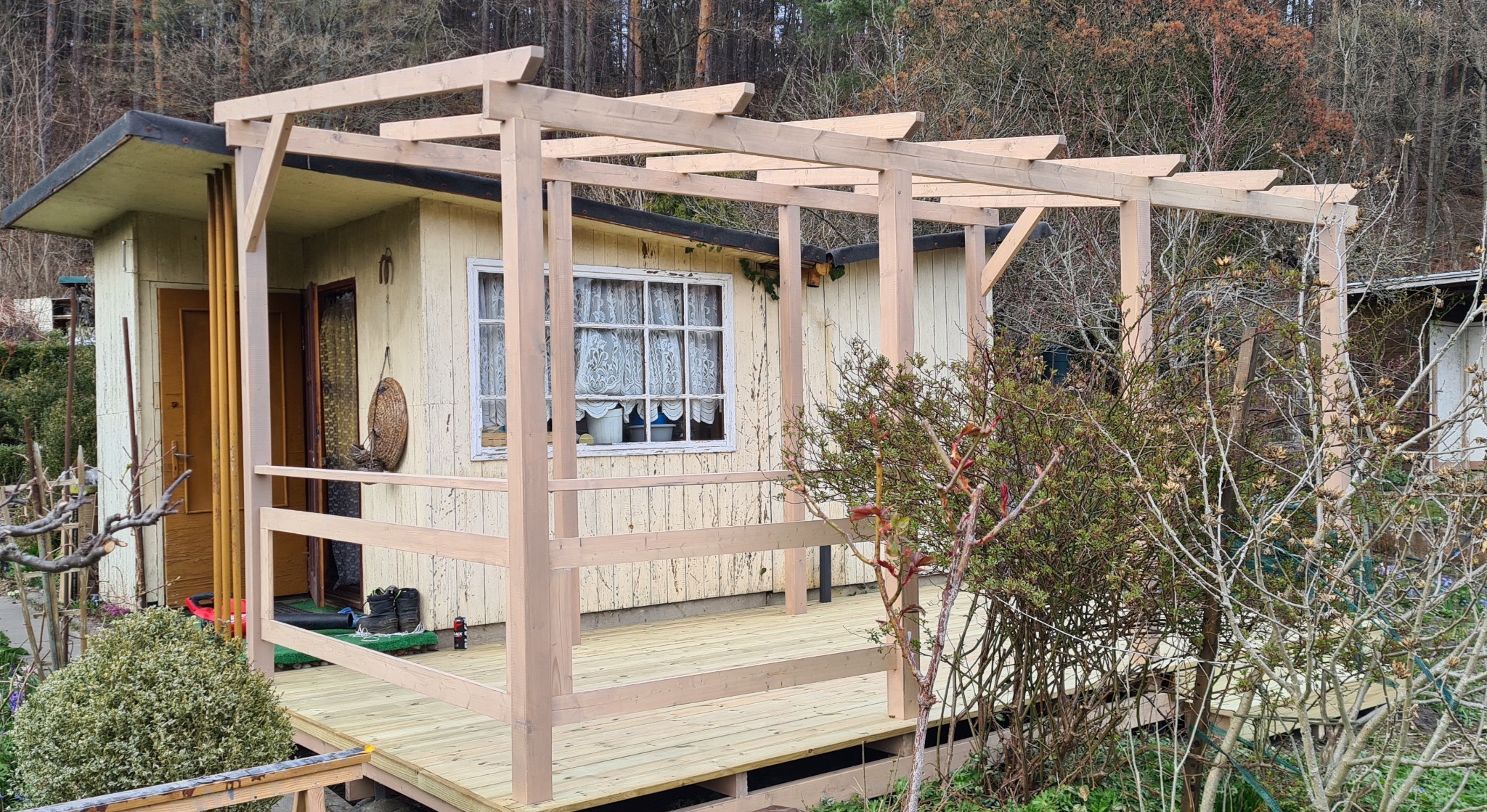 Drewniana konstrukcja zadaszenia nad tarasem przy domku, w trakcie budowy, widoczne elementy podtrzymujące i deski podłogowe, tło stanowią drzewa bez liści.