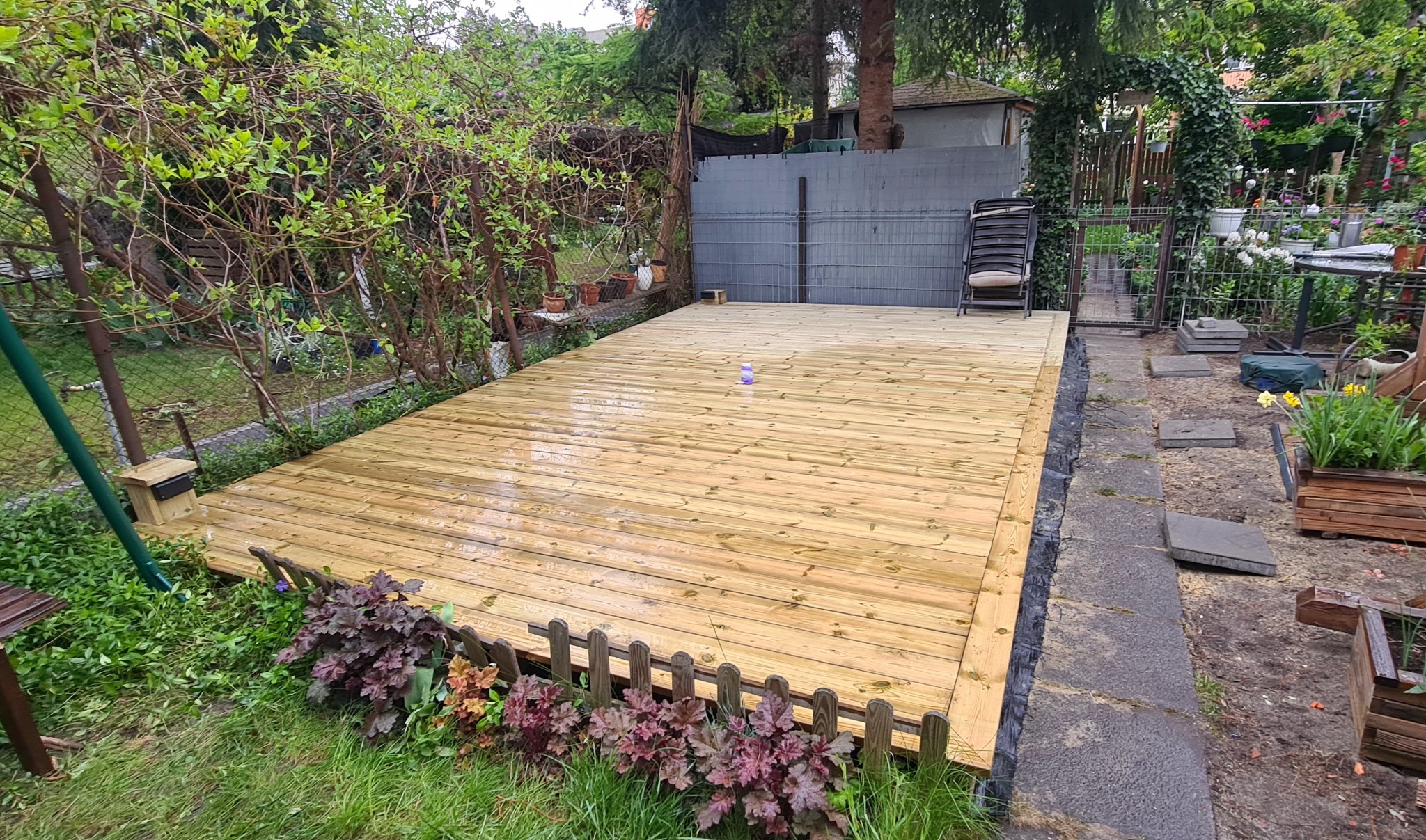 Świeżo położony taras z desek w ogrodzie, po deszczu, z widoczną fakturą drewna i elementami zieleni. W tle krzesło i metalowe ogrodzenie.