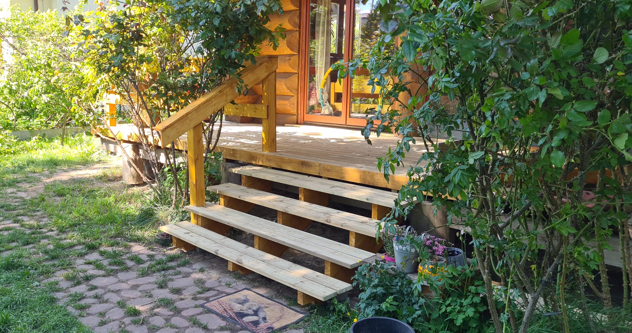 Drewniany taras z jasnego drewna z trzema schodami, otoczony zielenią. Wejście do domu z drewnianymi drzwiami. Kostka brukowa na ziemi.