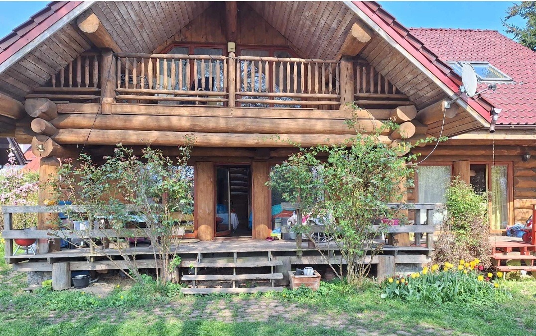 Drewniany dom z bali z tarasem i balkonem, czerwony dach z oknem dachowym i anteną satelitarną. Na tarasie roślinność, schody wejściowe. Dom w stylu rustykalnym.