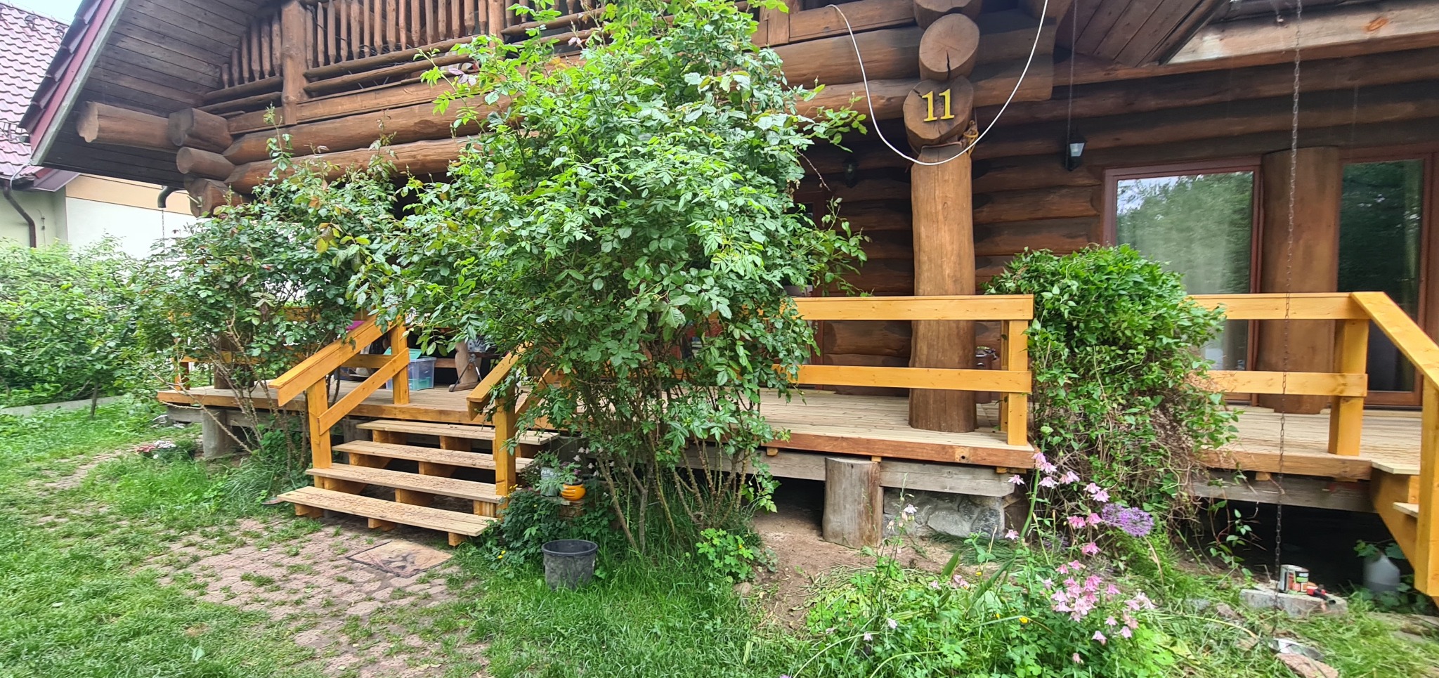 Drewniany taras z balustradą przy domu z bali, z roślinnością w tle. Schody prowadzą na taras, widoczne kwiaty i krzewy. Numer domu '11' na drewnianym filarze.