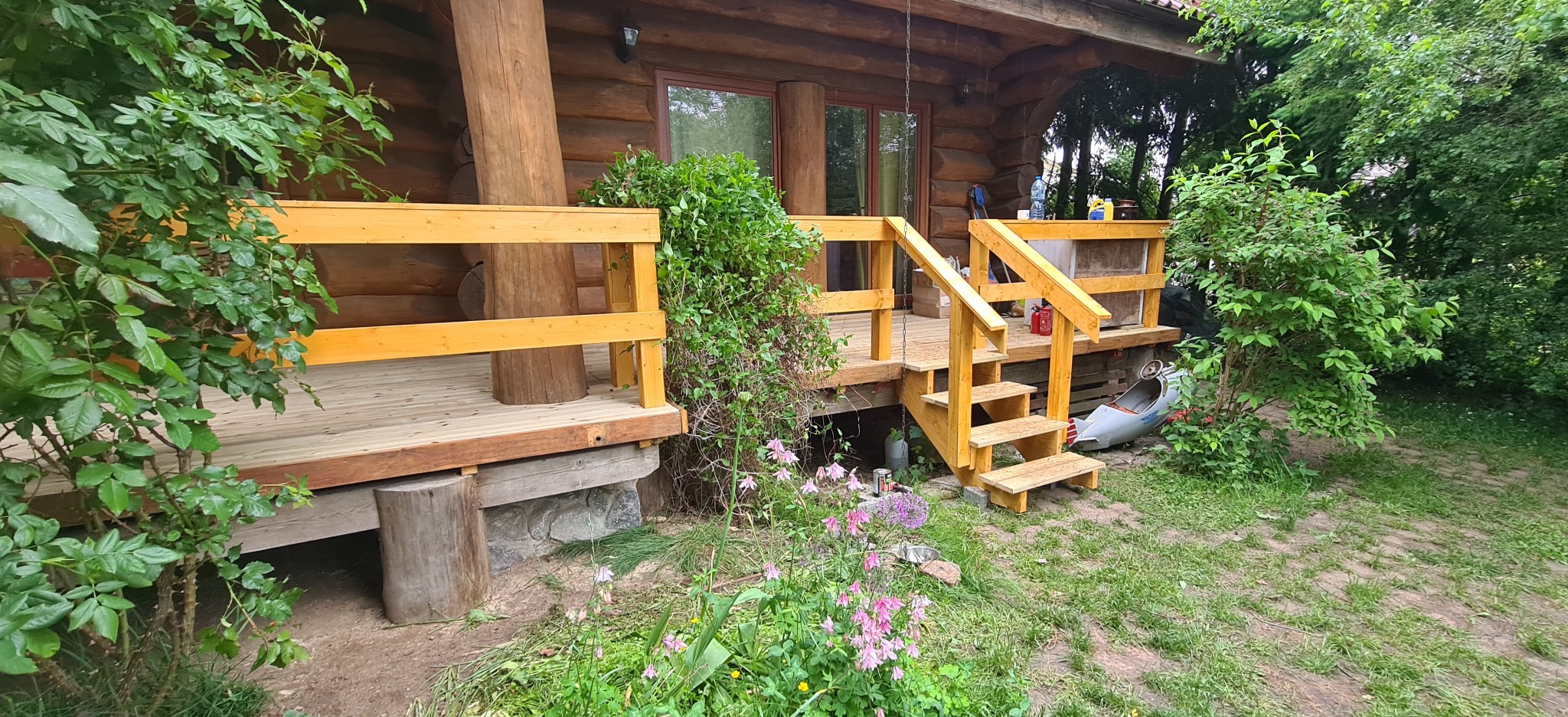 Drewniany taras przy domu z bali, z żółtymi barierkami i schodami. Elementy konstrukcyjne oparte na kamieniach. Bujna roślinność w tle i na pierwszym planie.