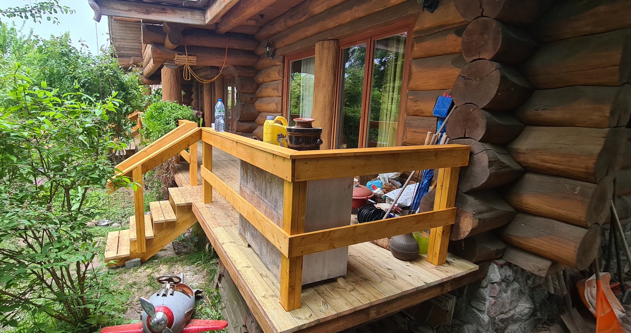 Drewniany taras przy domu z bali, widoczne schody i balustrada. Na tarasie ustawione donice i inne przedmioty. W tle drzewa i krzewy.