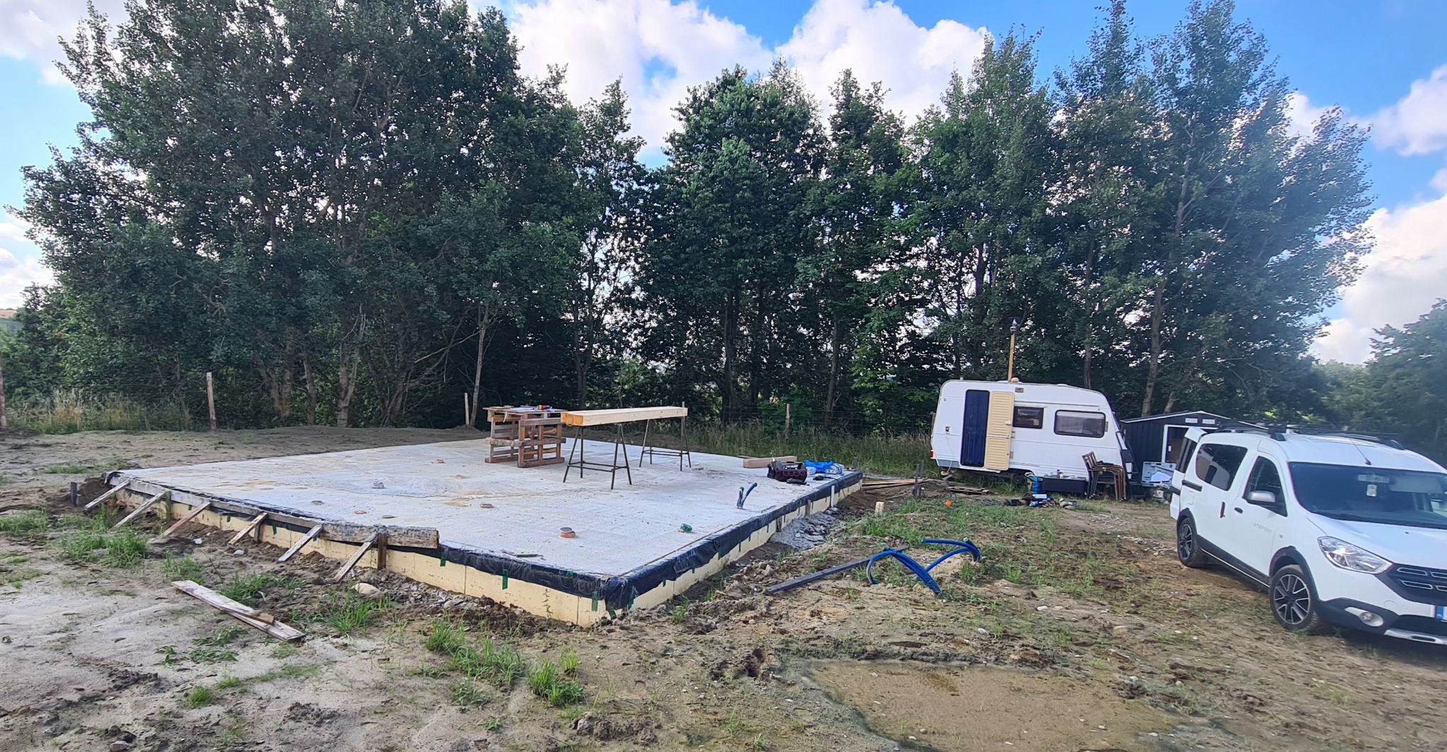 Betonowa płyta fundamentowa z izolacją, przygotowana pod budowę. W tle zieleń drzew, przyczepa kempingowa i biały samochód dostawczy.