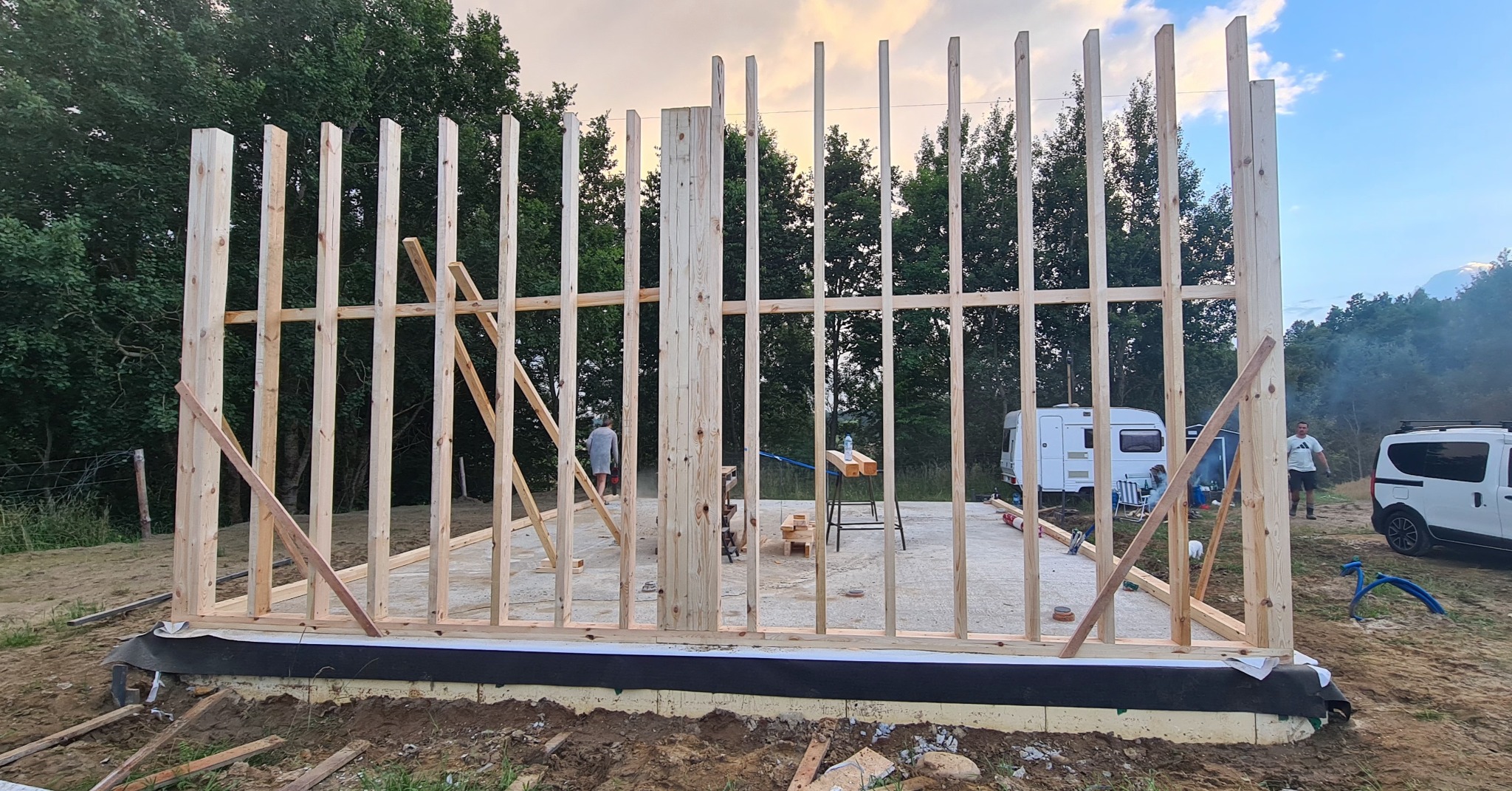 Surowa konstrukcja drewniana budynku w trakcie budowy, widoczne słupy i belki, na tle zalesionego terenu. W tle przyczepa kempingowa i samochód dostawczy.