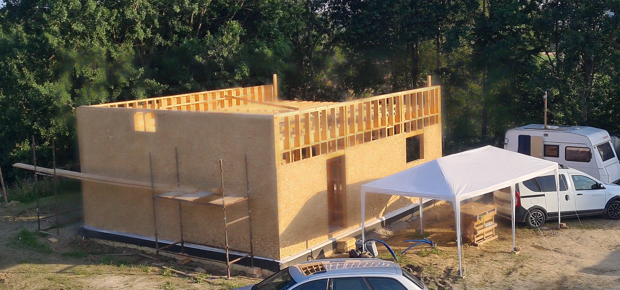 Budowa szkieletowej konstrukcji domu z drewna, widoczne ściany z płyt OSB, elementy więźby dachowej, na zewnątrz przyczepa kempingowa i biały namiot.