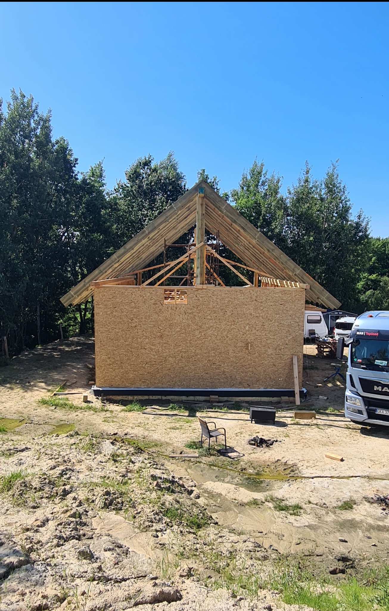 Budowa altany w lesie: konstrukcja z drewna i płyty OSB, widoczny szkielet dachu, drzewa w tle. Częściowo zabudowana ściana, teren budowy z piaskiem i błotem.