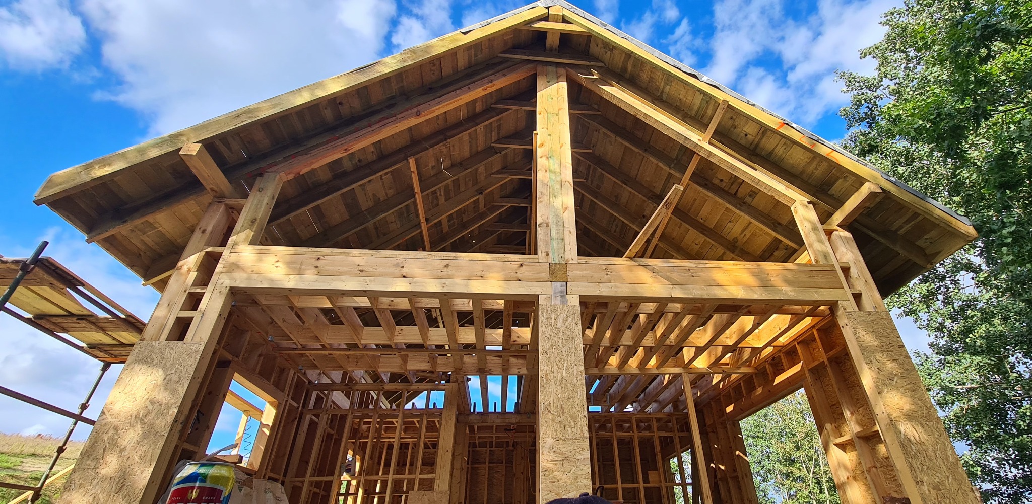 Drewniana konstrukcja budynku w trakcie budowy, widok z dołu na szkielet dachu i ścian, błękitne niebo w tle, widoczne elementy rusztowania i puszka po napoju.