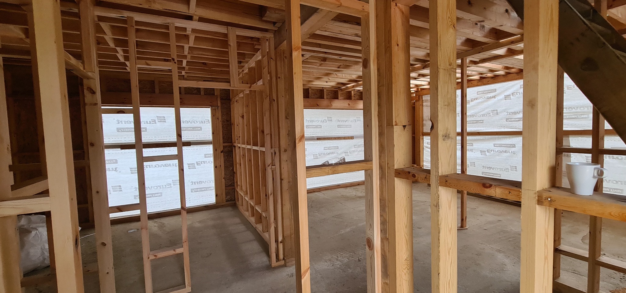 Surowa konstrukcja szkieletowa budynku z drewnianych belek, widok z wnętrza, z oknami zabezpieczonymi folią i kubkiem na parapecie. Etap budowy domu.