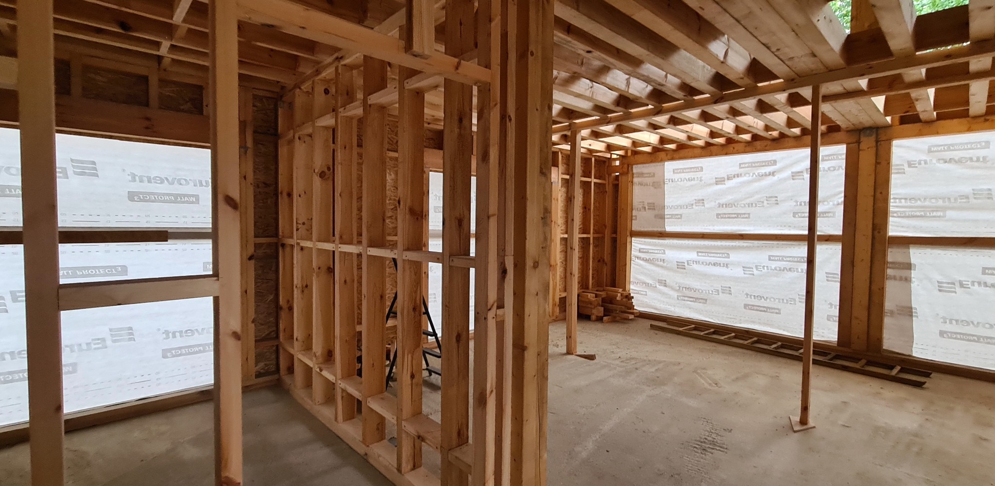 Surowa konstrukcja z drewna, tworząca ramy ścian i stropu, widoczna od wewnątrz, z folią ochronną na zewnątrz. Etap budowy domu z widocznymi elementami konstrukcyjnymi.