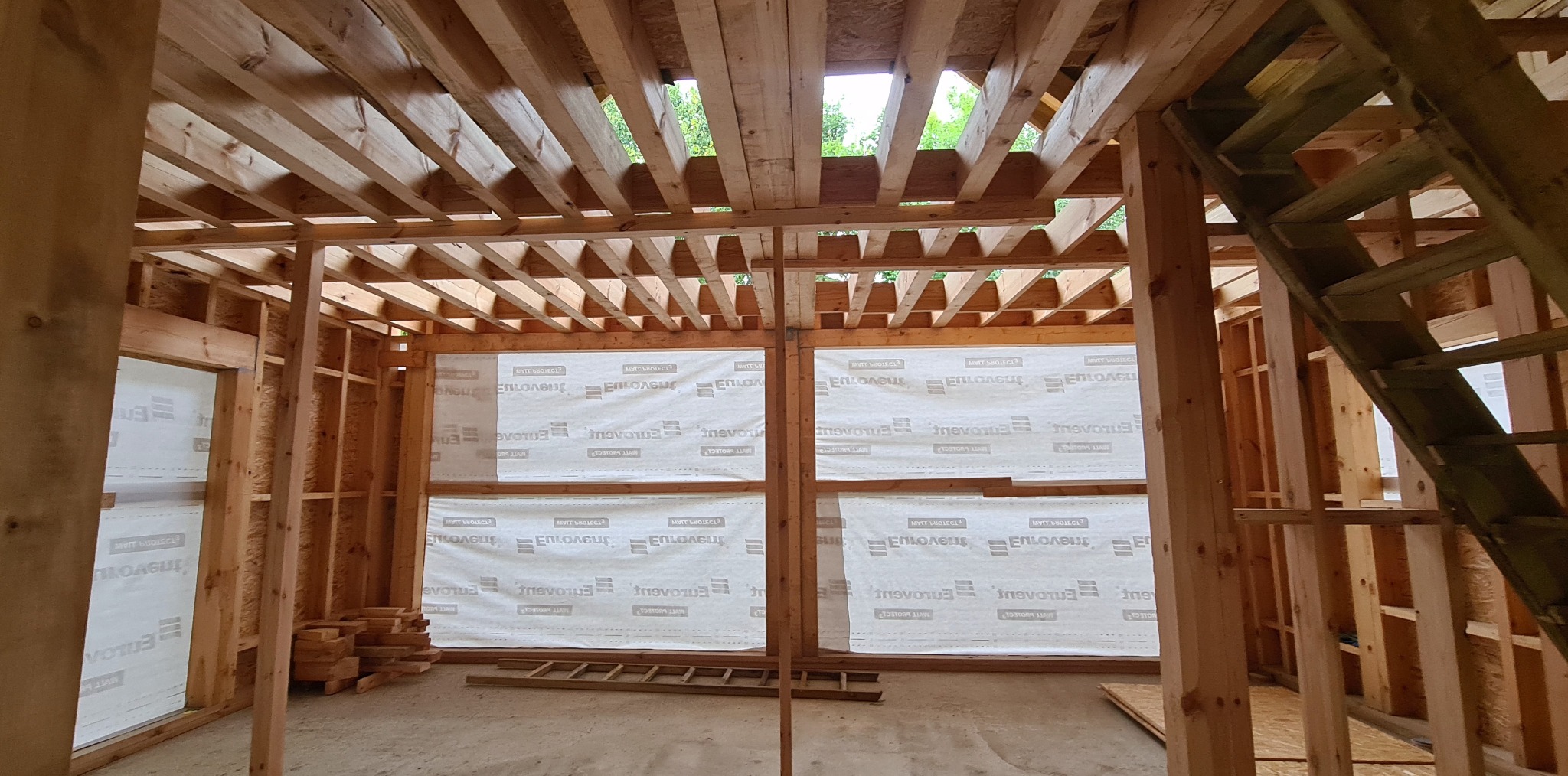 Konstrukcja drewniana w trakcie budowy: szkielet ścian z membraną, widoczna więźba dachowa, częściowo wykonane schody. Wnętrze w stanie surowym, przygotowane do dalszych prac.