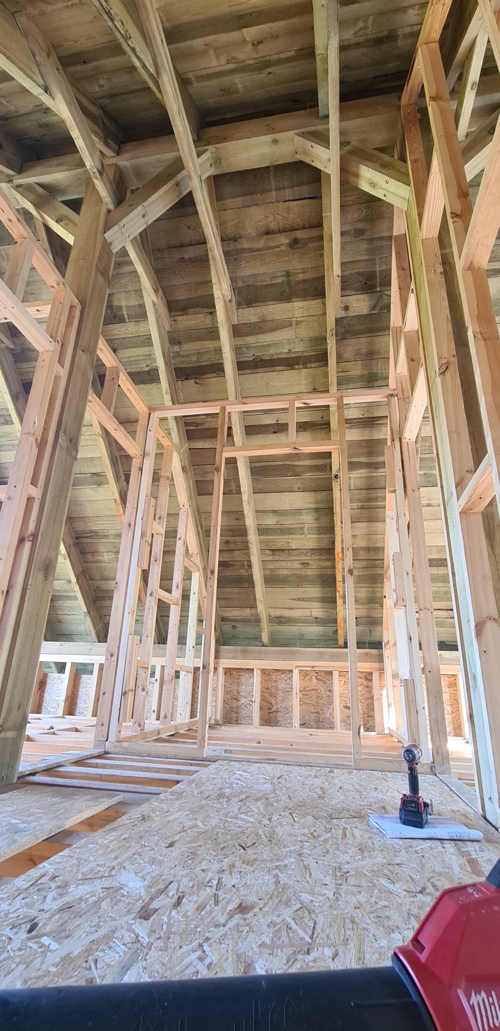 Konstrukcja drewniana w trakcie budowy, widok z dołu na szkielet dachu i ścian. Na podłodze wkrętarka i notatnik. Surowe drewno, jasne oświetlenie.