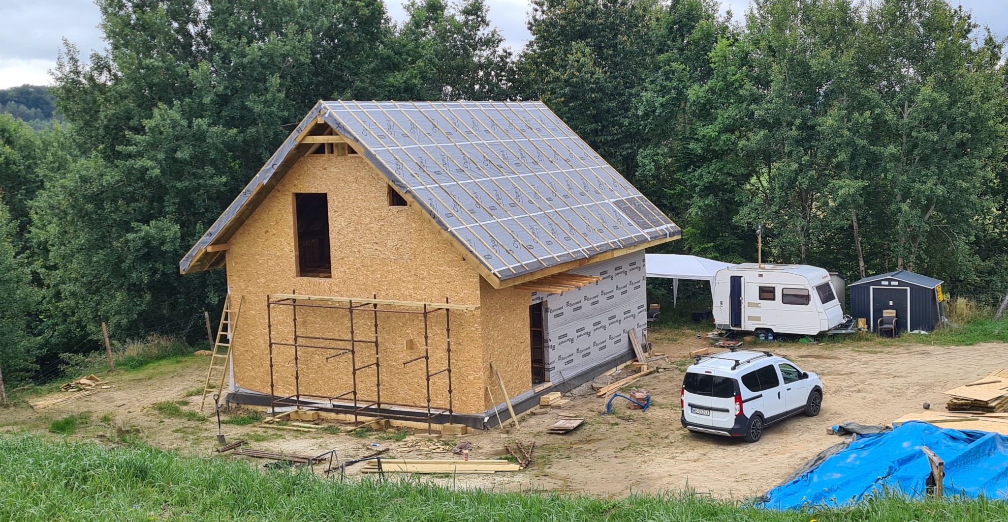 Surowy stan domu w budowie z deskami OSB, widoczne rusztowania, dach z membraną, przyczepa kempingowa i auto na działce, otoczenie zielone.
