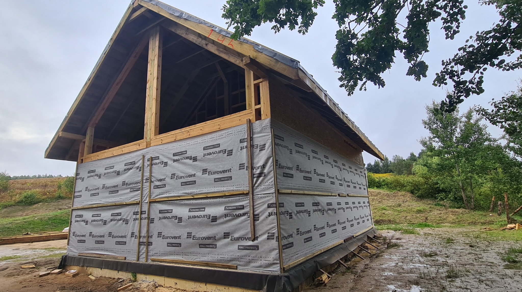 Budowa drewnianej konstrukcji domu w trakcie wznoszenia, z widoczną membraną Eurovent na ścianach i otwartą konstrukcją dachu, na tle zieleni i pochmurnego nieba.