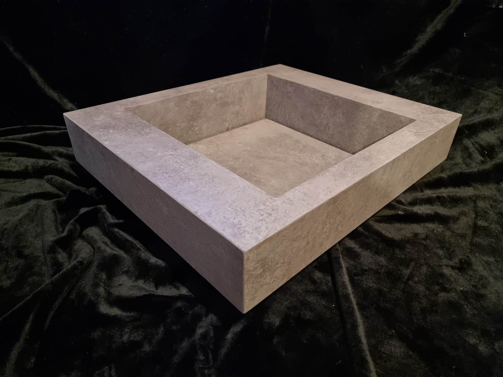 Kwadratowa umywalka z szarego kamienia kompozytowego na tle czarnego aksamitu, widok z góry pod kątem.