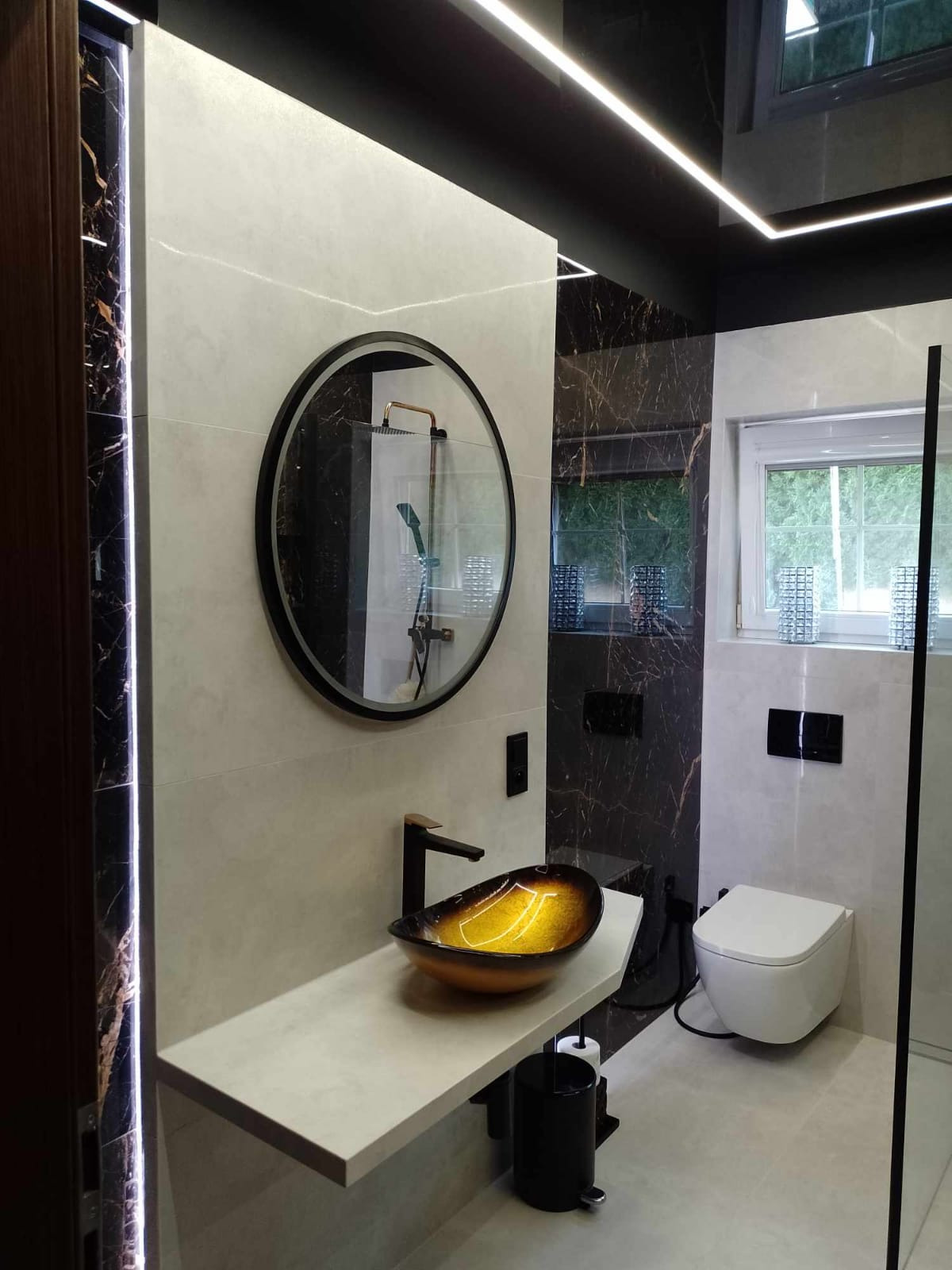 Nowoczesna łazienka z umywalką w kształcie łódki w odcieniach złota i czerni, okrągłe lustro z czarną ramą, płytki imitujące marmur i podświetlenie LED wzdłuż krawędzi.