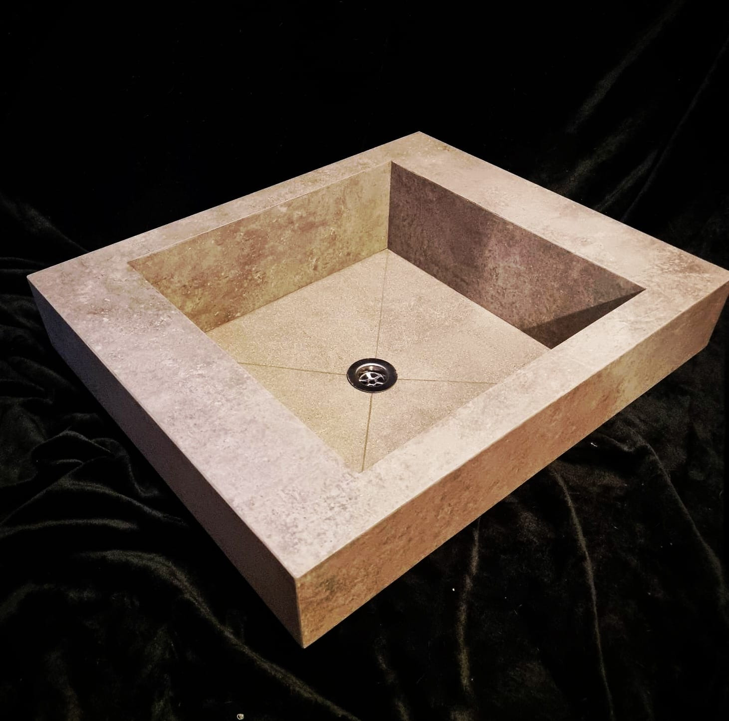 Kwadratowa umywalka z beżowego kamienia z delikatną fakturą, widoczny odpływ i cztery linie wyznaczające spadek w kierunku odpływu, ustawiona na czarnym, aksamitnym tle.