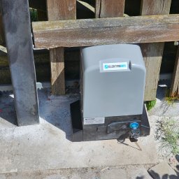 DG-TECH Gabriela Jarmasz - Szary automat do bramy z kluczem w zamku, zamontowany przy drewnianym ogrodzeniu z widoczną listwą zębatą.