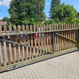 DG-TECH Gabriela Jarmasz - Otwarta, drewniana brama przesuwna z widocznym mechanizmem automatycznym, na tle kostki brukowej i zieleni.