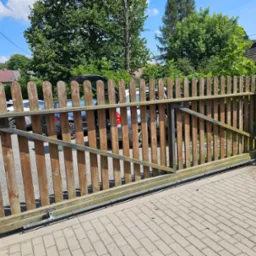 Otwarta, drewniana brama przesuwna z widocznym mechanizmem automatycznym, na tle kostki brukowej i zieleni.