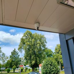 DG-TECH Gabriela Jarmasz - Kopułkowa kamera monitoringu zamontowana pod zadaszeniem, widok na drzewa i zaparkowane samochody w tle.