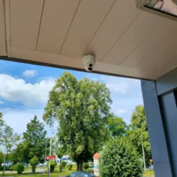 Kopułkowa kamera monitoringu zamontowana pod zadaszeniem, widok na drzewa i zaparkowane samochody w tle.