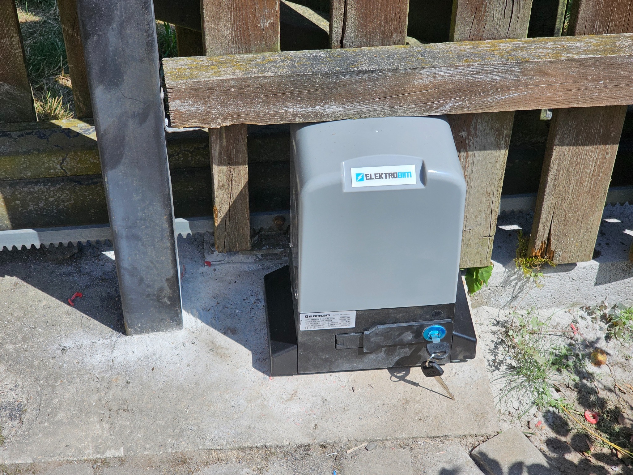 Szary automat do bramy z kluczem w zamku, zamontowany przy drewnianym ogrodzeniu z widoczną listwą zębatą.