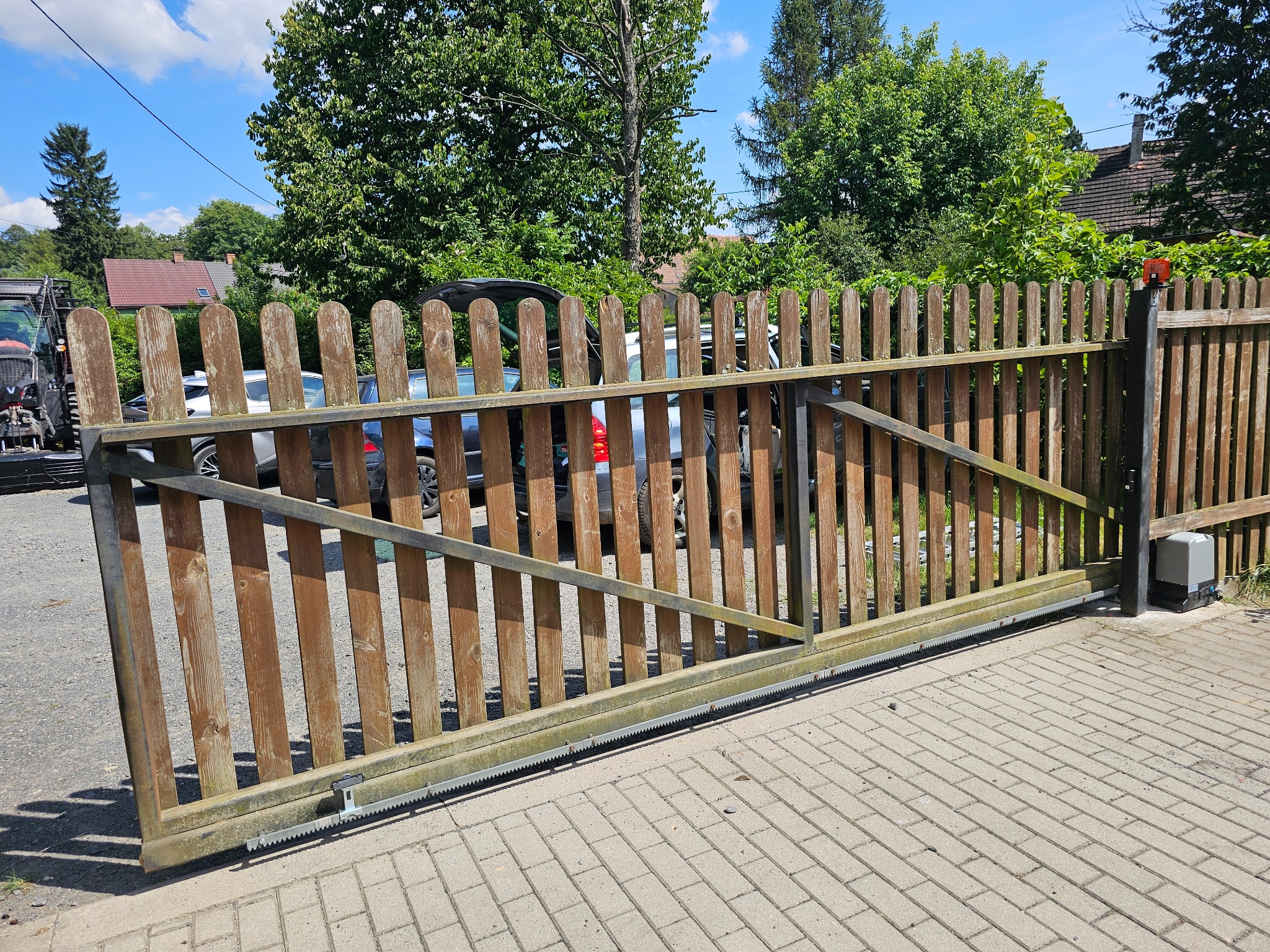 Otwarta, drewniana brama przesuwna z widocznym mechanizmem automatycznym, na tle kostki brukowej i zieleni.