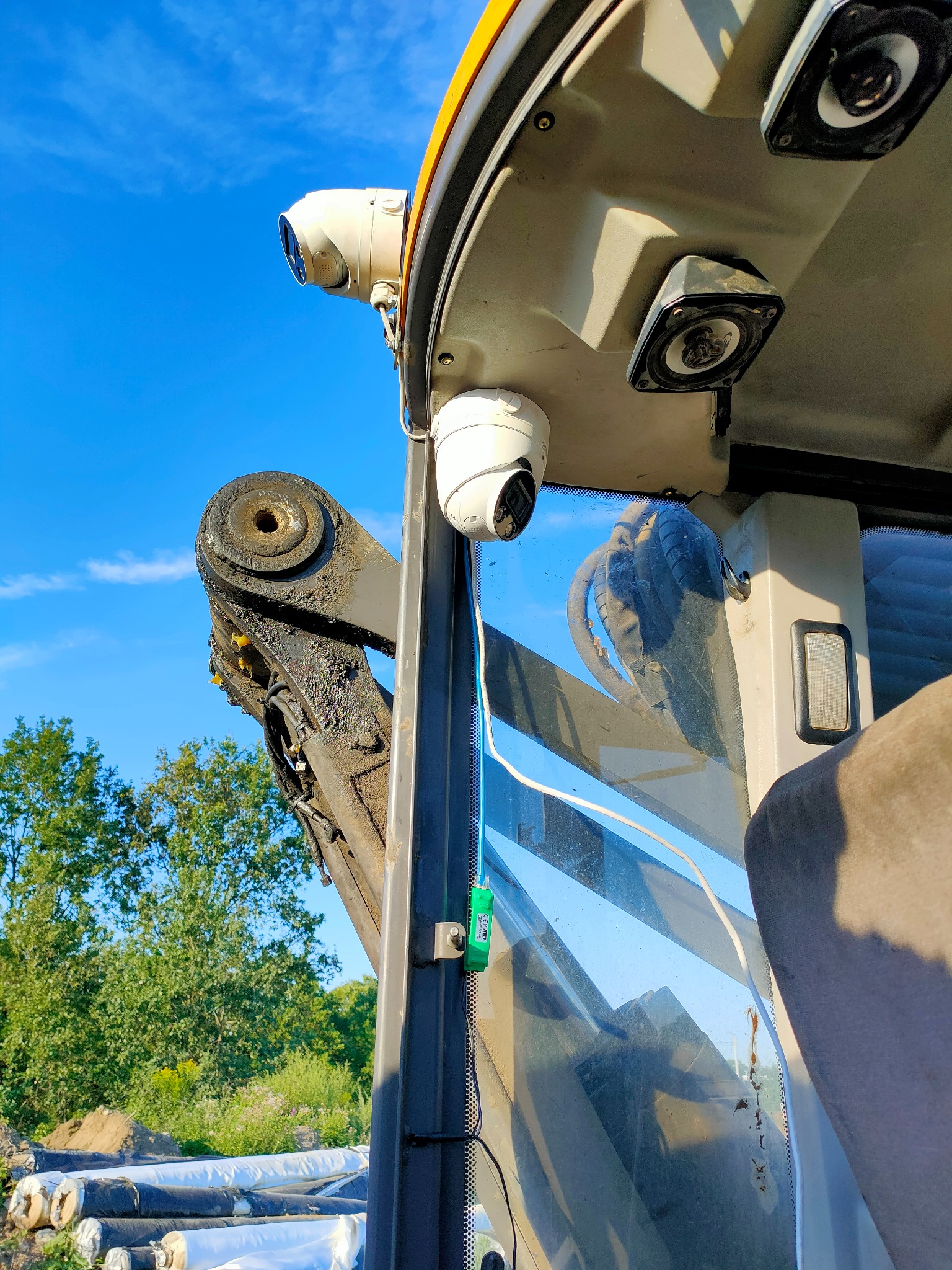 Wnętrze kabiny maszyny budowlanej z zamontowaną kamerą monitoringu na suficie i dodatkowym oświetleniem, widok na ramię maszyny i błękitne niebo.